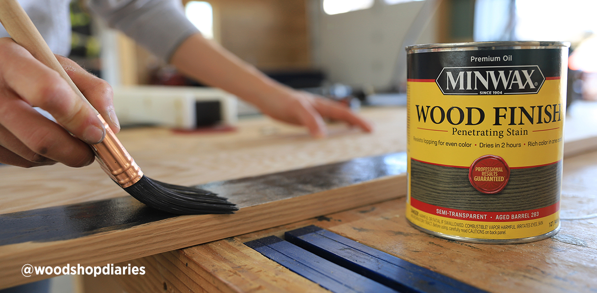 A DIYer in her garage stains a piece of wood using a paint brush and a one quart can of Minwax Wood Finish Aged Barrel.